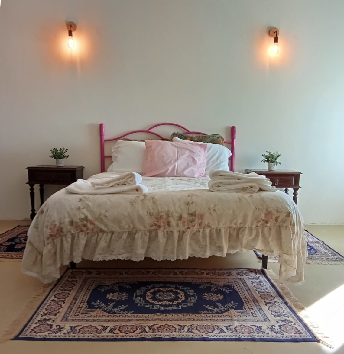 Detalhe do Quarto de casal: vista de frente da cama, com mesinhas de cabeceira, tapetes com motivos de flores e candeeiros de parede