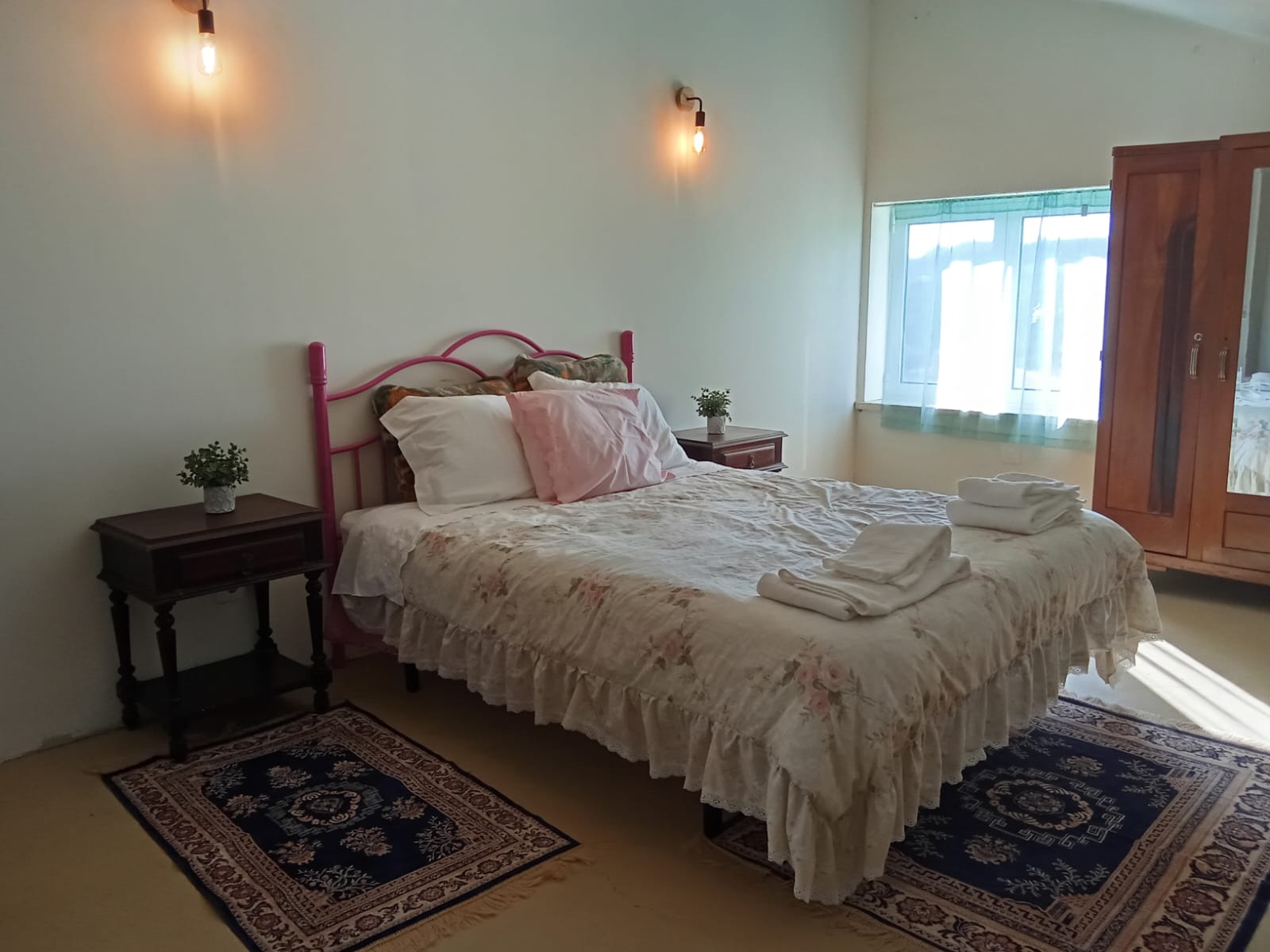 Detalhe do Quarto de casal: cama, com mesinhas de cabeceira, tapetes com motivos de flores, candeeiros de parede, e a janela com estores el�ctricos